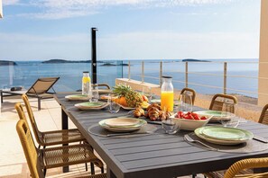 Enjoy a meal with a view of the sea from this outdoor dining area, featuring a table set with plates, cutlery, and fresh fruits.