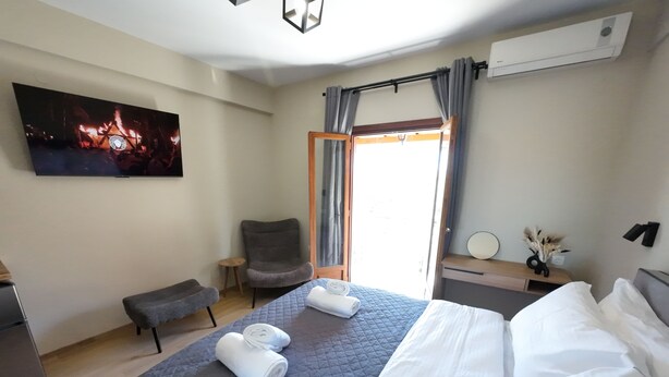 A hotel room with a bed, a chair, a small table, a TV, and a window with curtains. - Nel Paese Room (Skiathos)