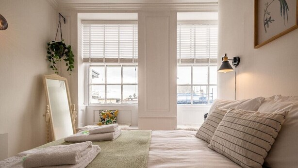 A bedroom with a bed, a bedside table with towels, a mirror, and a window with blinds. - Stylish and bright City Centre apartment (Exeter)