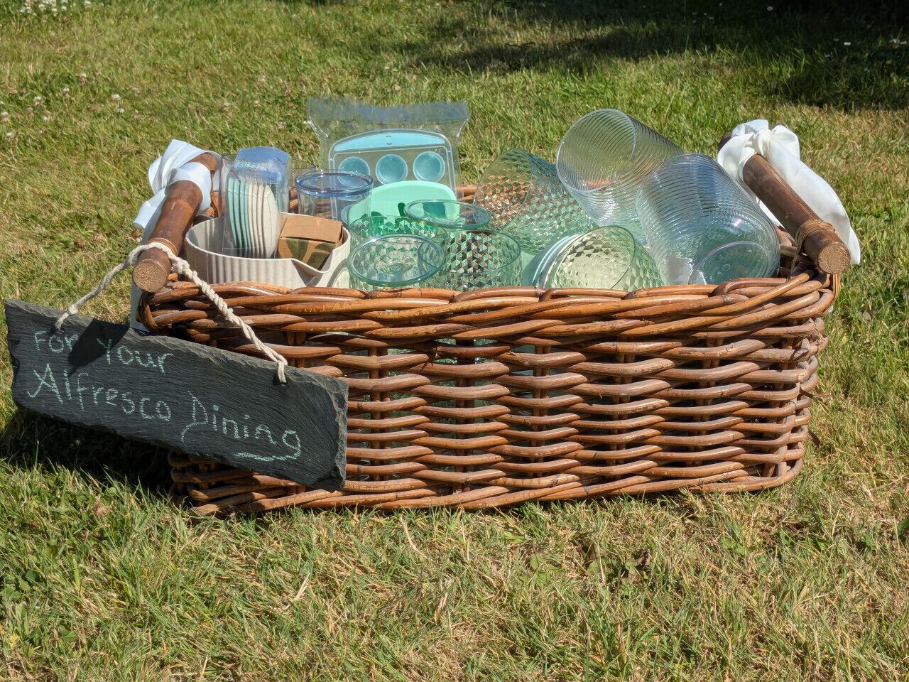 Un panier en osier rempli de divers objets en verre et une pancarte sur laquelle on peut lire : « Pour vos repas en plein air ».