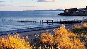 A coastal scene with a wooden pier extending into calm waters, a grassy foreground, and a distant shoreline with buildings. - Charming character lodge in cemetery grounds (Devon)