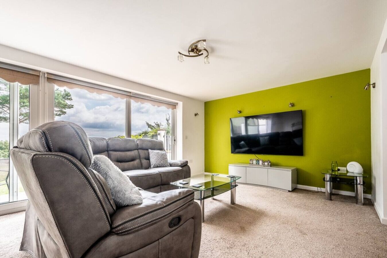 A modern living room with a grey leather sofa, a glass coffee table, and a large flat-screen TV mounted on a green wall.