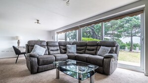 A modern living room with a sectional sofa, a glass coffee table, and large windows offering a view of the outdoors. - 2 BR Seaside Flat/ Parks 2 - Garden & Ocean Views (Dawlish)