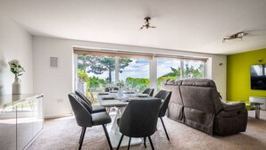 A modern living room with a dining area, large windows, a ceiling fan, and a green accent wall. - 2 BR Seaside Flat/ Parks 2 - Garden & Ocean Views (Dawlish)