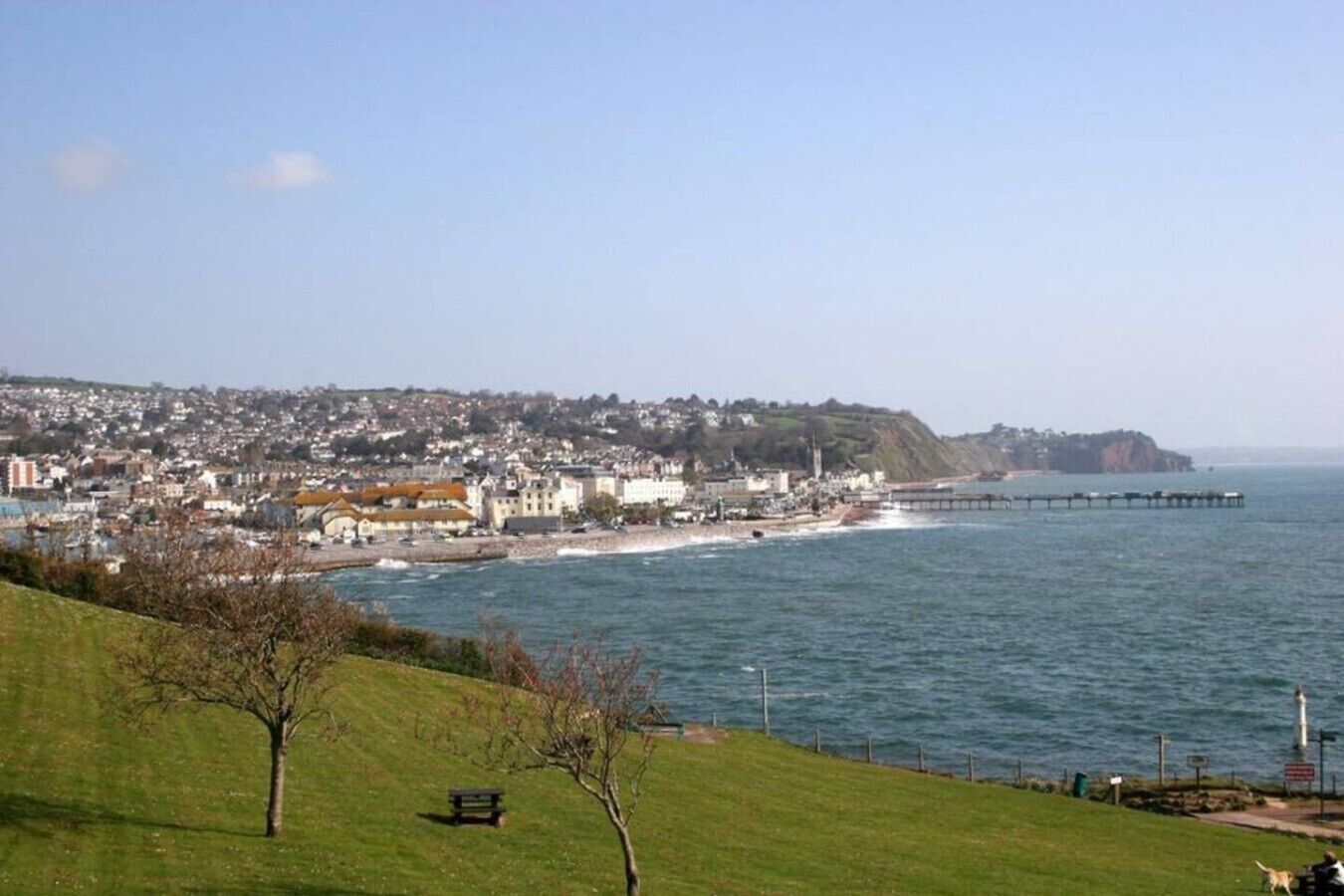 A coastal town with a pier extending into the sea, a grassy hill with a bench, and a clear sky.