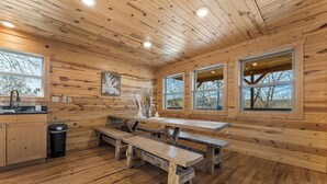 A wooden cabin interior with a kitchen area, wooden benches, and large windows.