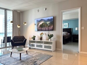 Ein modernes Wohnzimmer mit Sofa, Couchtisch, Wand-Fernseher und Blick durch eine offene Tür in ein Schlafzimmer.