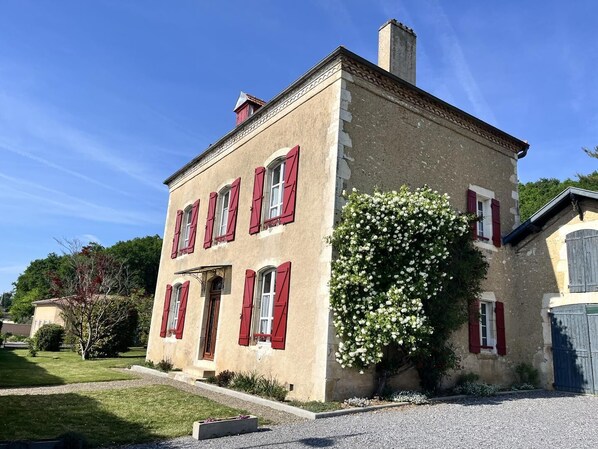 A two-story building with red shutters, a chimney, and a garden with white flowers.