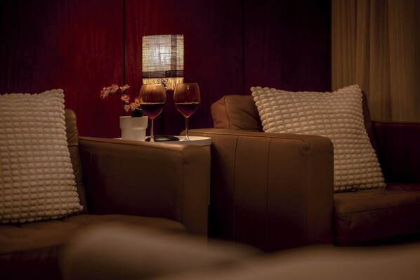 A dimly lit room with a brown leather sofa, two white textured pillows, a small white table with a vase of pink flowers and two wine glasses. - Sintra Dream Garden Villa (Sintra)