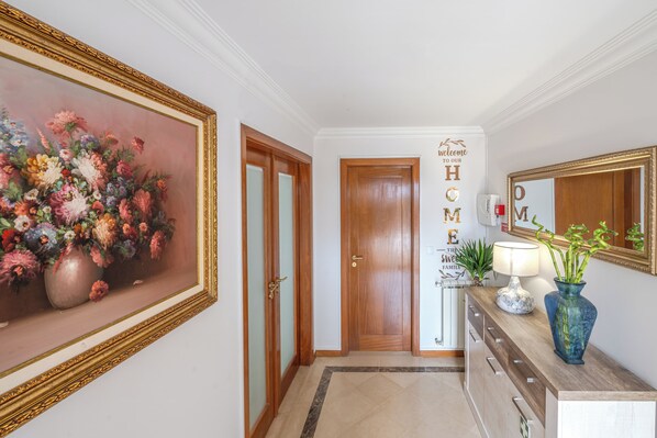 Un couloir orné d’un tableau représentant des fleurs, avec une porte en bois, un panneau d’accueil, un miroir et un vase contenant des plantes.