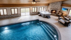 An indoor swimming pool with a hot tub, a fireplace, and a seating area with a view of the outdoors.