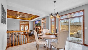 A dining area with a round table, chairs, and a chandelier. There is a view of the outdoors through large windows.