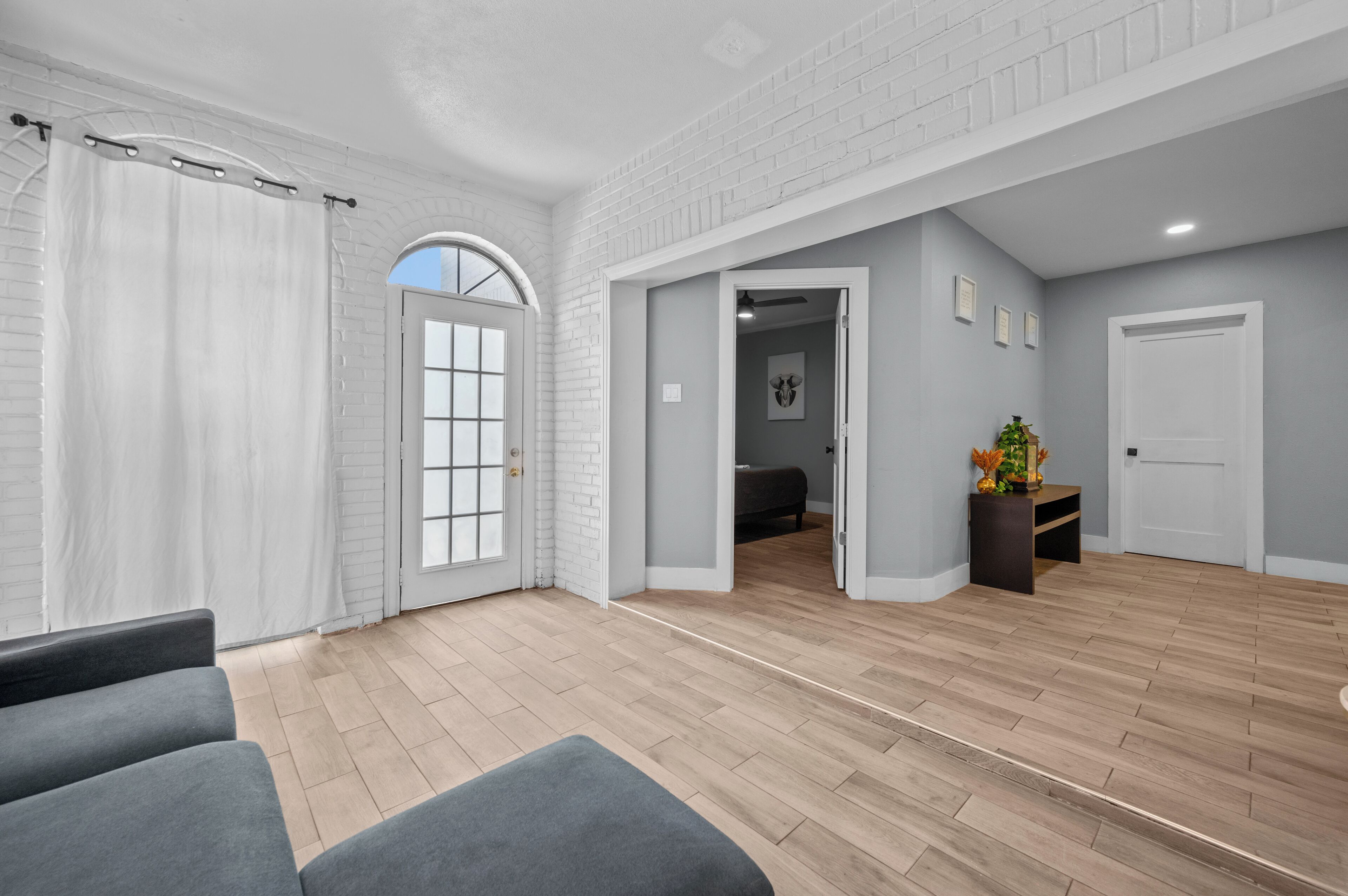 Un salon spacieux avec une grande fenêtre, une porte donnant sur une autre pièce et une petite table sur laquelle est posée une plante.