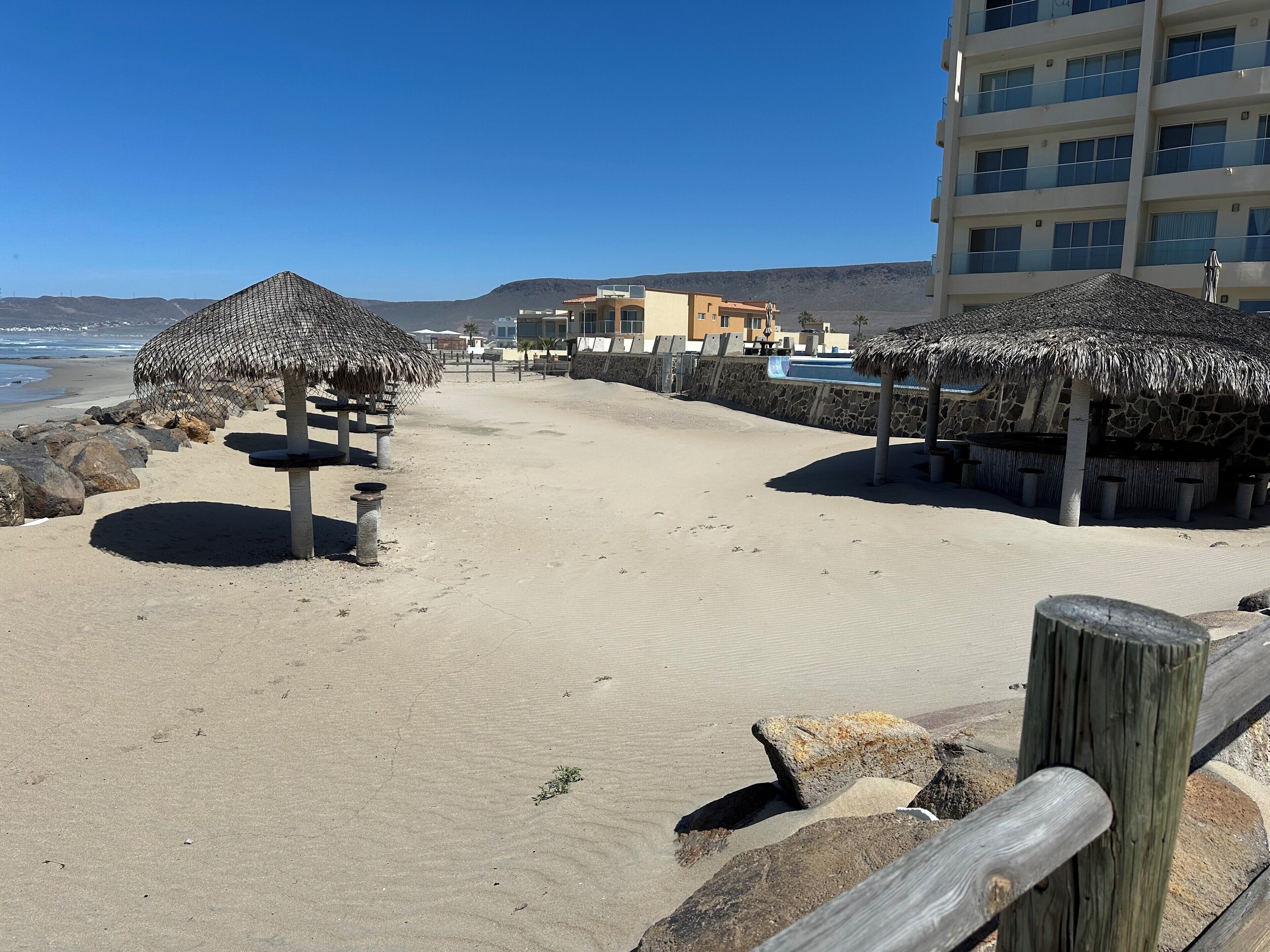 Een strand met rieten parasols, een zandpad, een houten hek en een gebouw op de achtergrond.
