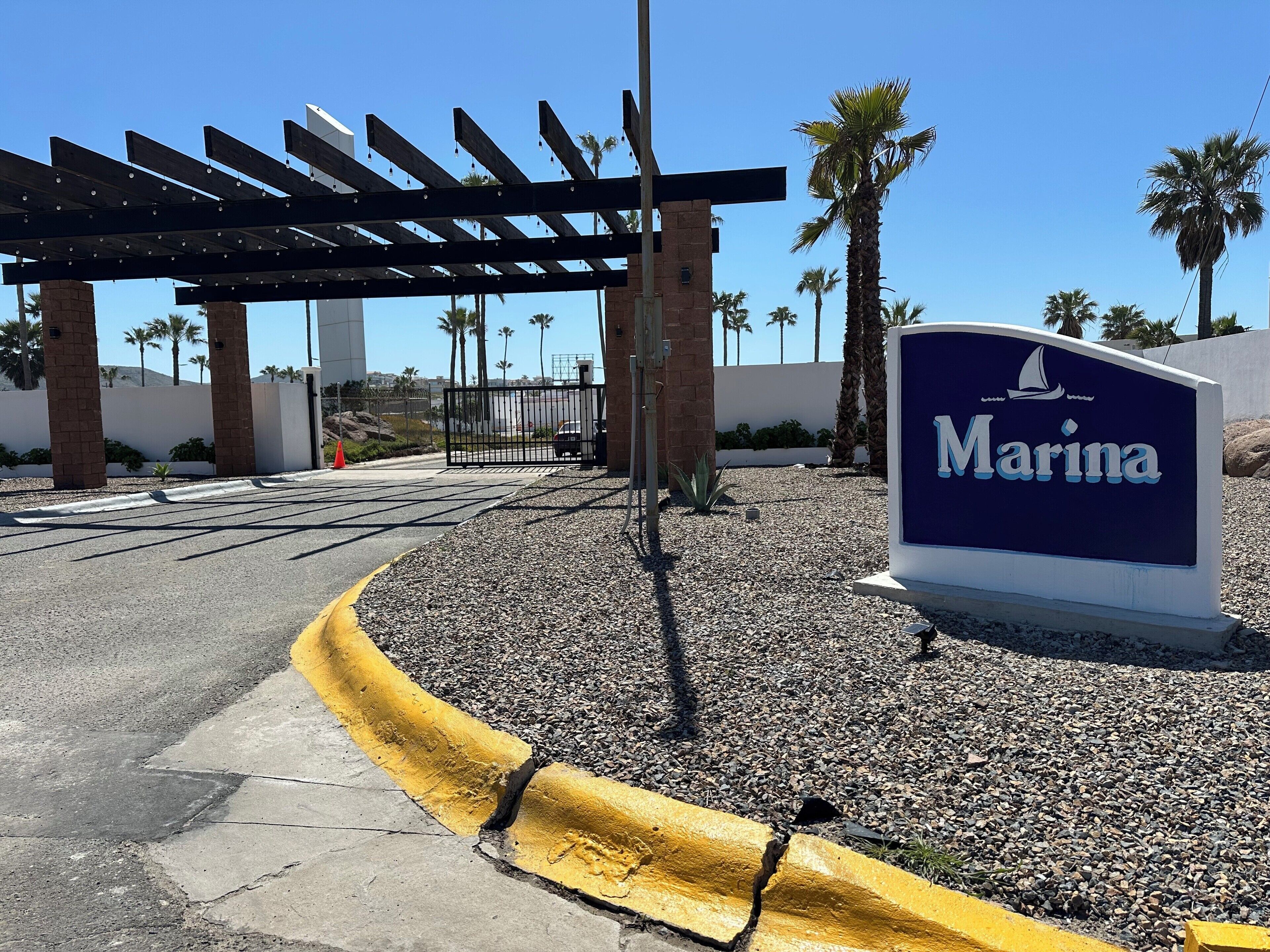 A marina entrance with a sign, a pergola, palm trees, and a clear sky.