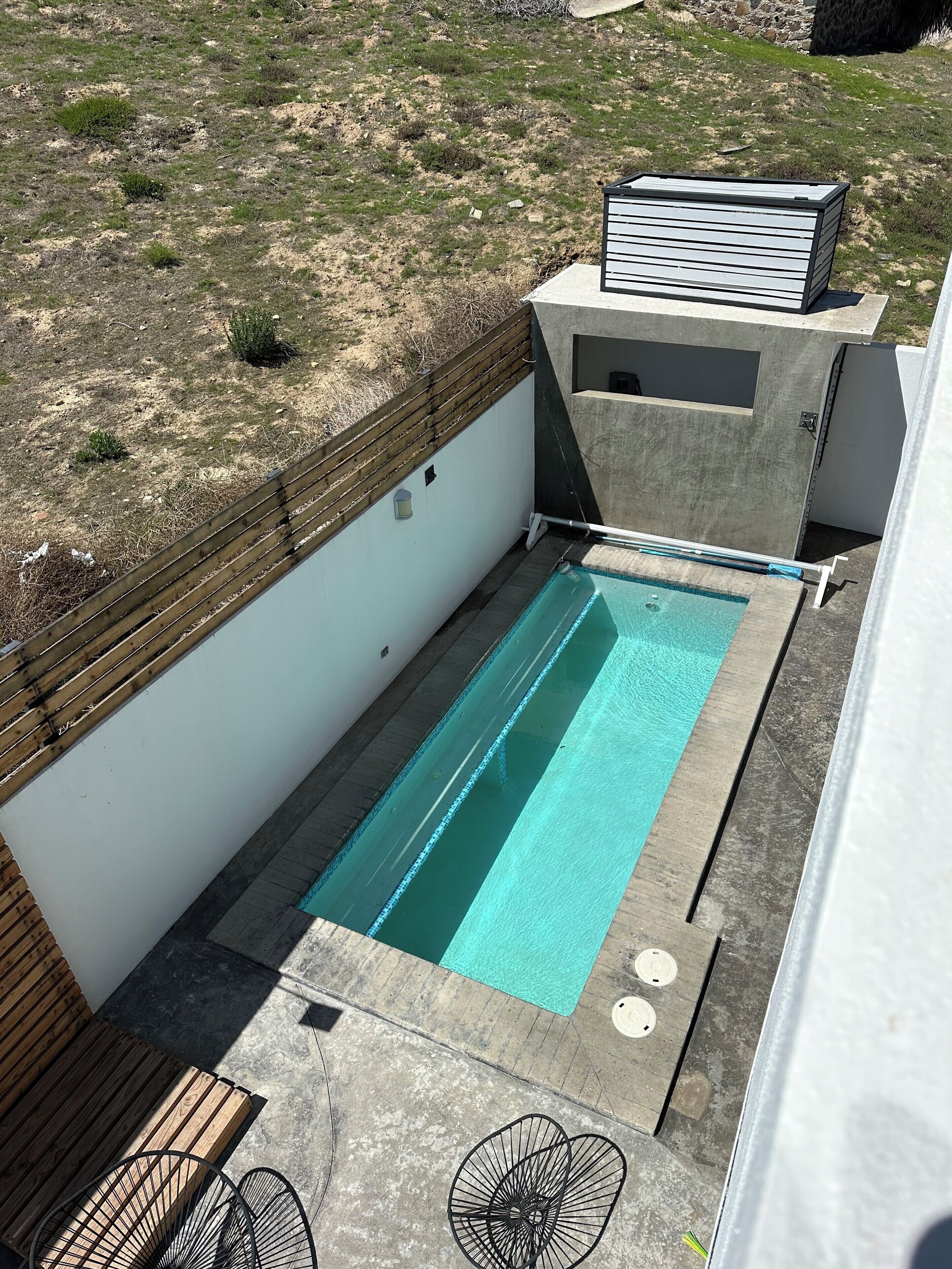 A rectangular pool with a wooden deck and a concrete border, surrounded by a white wall and a wooden fence.