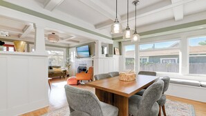 Un comedor con una mesa de madera y sillas grises, un televisor al fondo y vistas al exterior a través de amplios ventanales.