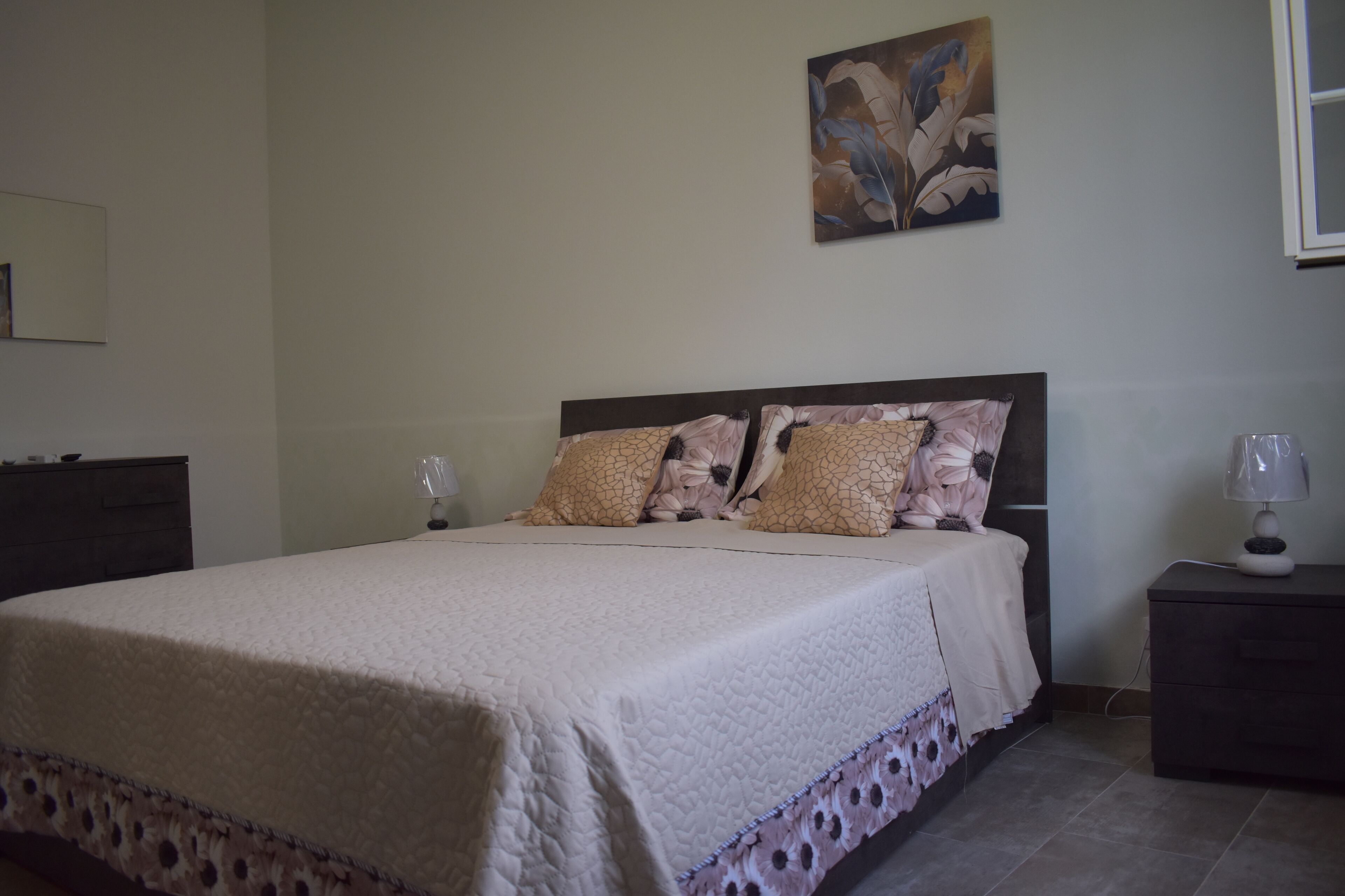 Ein ordentlich bezogenes Bett mit einem blumigem Überwurf und Kissen, ein Nachttisch mit einer Lampe und ein Bild an der Wand.