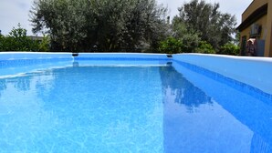Una piscina azul cristalina con borde de azulejos, rodeada de follaje y un edificio al fondo.