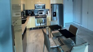 A modern kitchen with stainless steel appliances, a dining area with a glass table and chairs, and a view of the adjacent room.
