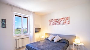 Ett sovrum med en säng, två sänglampor, ett element, ett fönster med gardiner och ett konstverk som hänger på väggen.