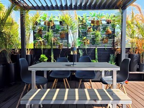 A covered outdoor dining area with a table and chairs, surrounded by potted plants and a wooden deck.