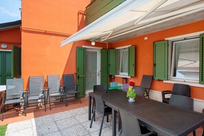 Um pátio externo com mesa e cadeiras, persianas verdes e toldo branco.