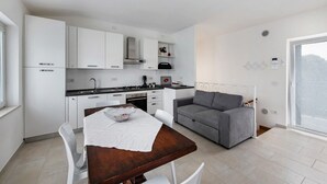 Una cocina moderna con armarios blancos, una mesa de comedor con sillas, un sofá gris y un ventanal amplio.