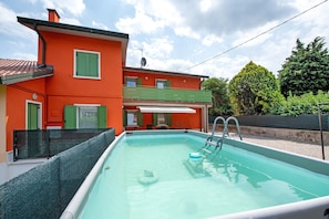 A swimming pool with a ladder, surrounded by a house with green shutters and a red facade.