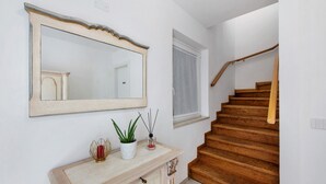 Una escalera con escalones y barandilla de madera, una pared blanca con un espejo grande, y una pequeña planta sobre una consola de madera.