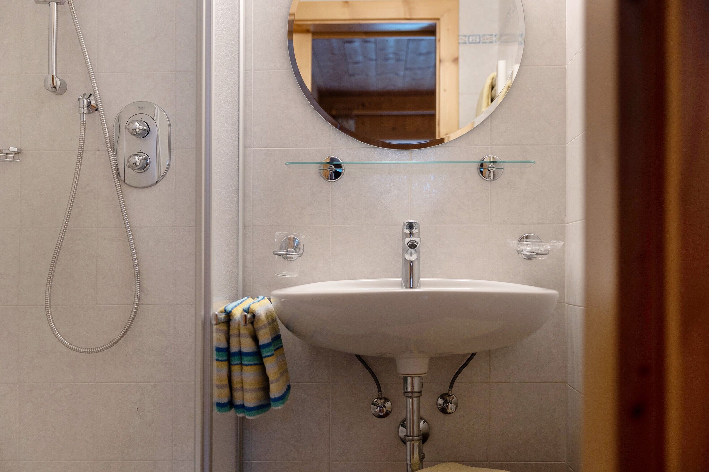 Une salle de bain avec un lavabo blanc, une étagère en verre au-dessus, un miroir rond et une douche équipée d’un pommeau de douche à main.