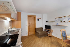 A compact kitchen with a sink, stove, and wooden cabinets. There is a wooden door, a television on a stand, and a bulletin board on the wall. - Apartment 'Almrausch' with Mountain View, Shared Terrace and Wi-Fi (St. Magdalena gsies)