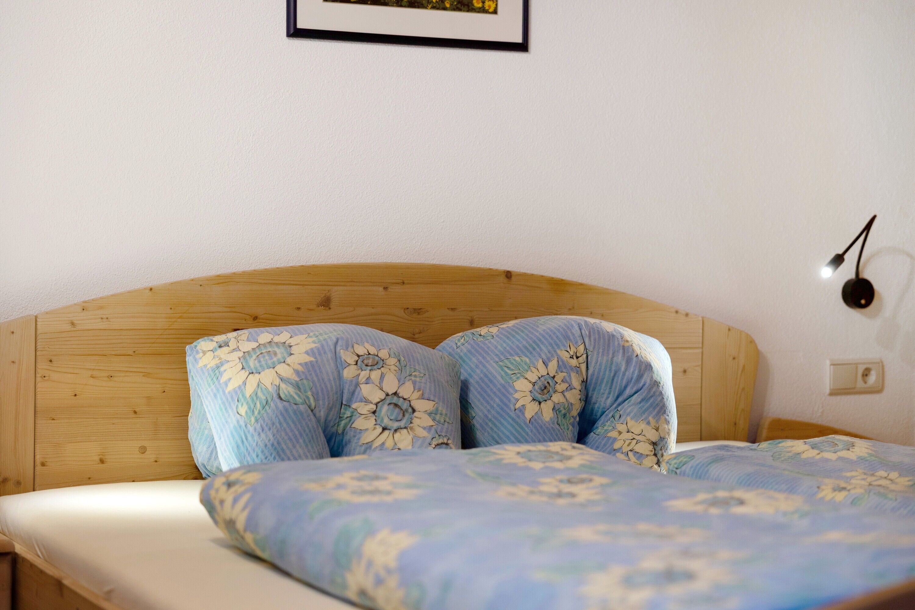 A bed with floral bedding, a wooden headboard, a wall-mounted light, and a framed picture above the bed.