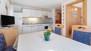 Uma cozinha compacta com armários brancos, uma mesa de jantar com uma cadeira azul e uma televisão sobre um suporte de madeira.