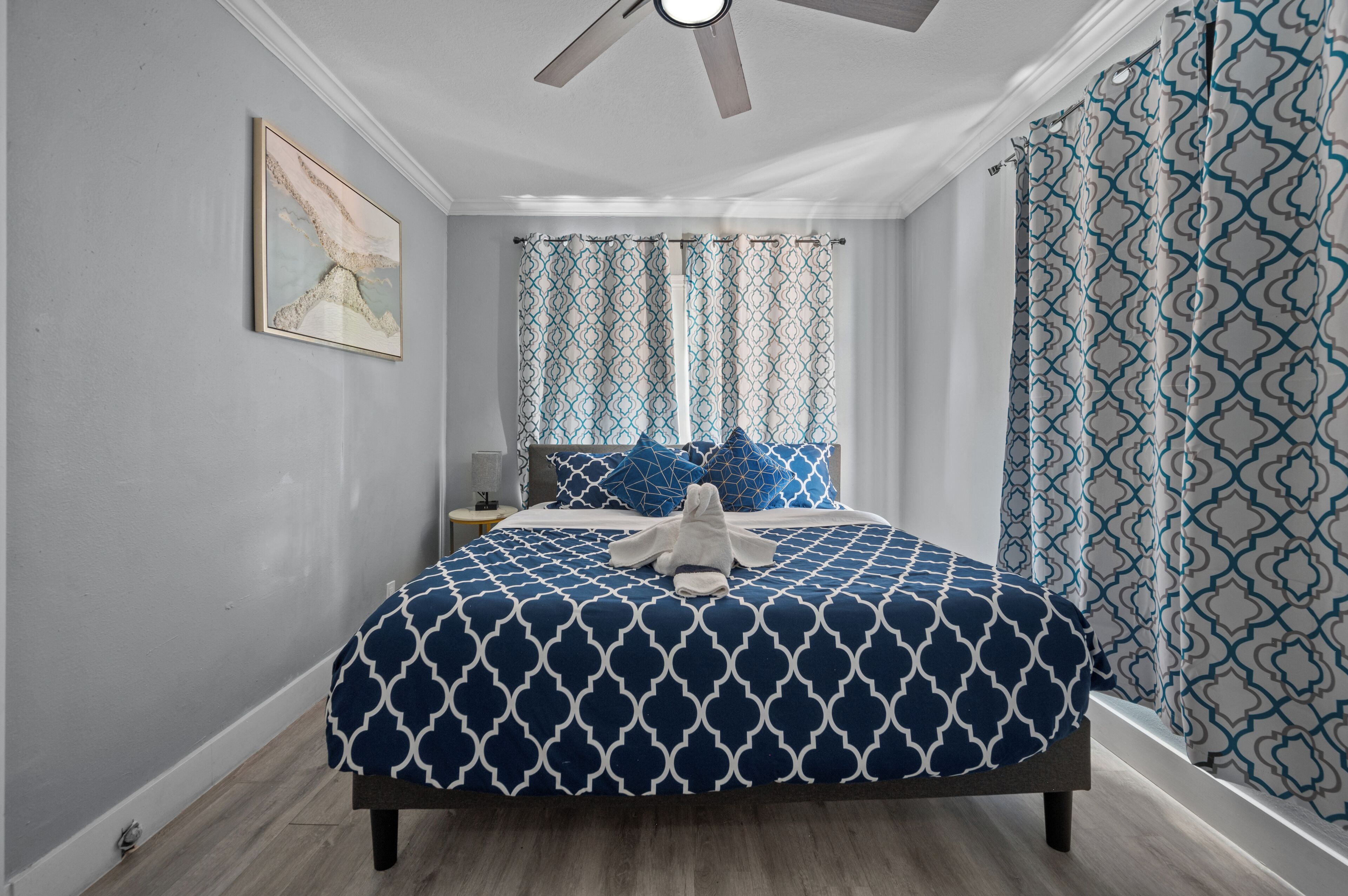 Un dormitorio con cama, ventilador de techo, cortinas estampadas y un cuadro en la pared.