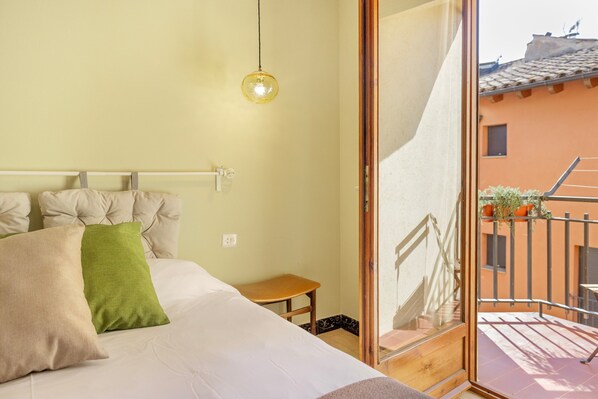 A bedroom with a bed, pillows, a bedside table, a hanging light fixture, and a view of an outdoor balcony with a railing and potted plants.