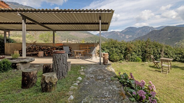Un espacio al aire libre techado con vista a montañas, una mesa de madera, sillas y un hogar.