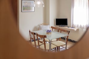 A dining area with a glass table, wooden chairs, a remote control, and a television on a stand.