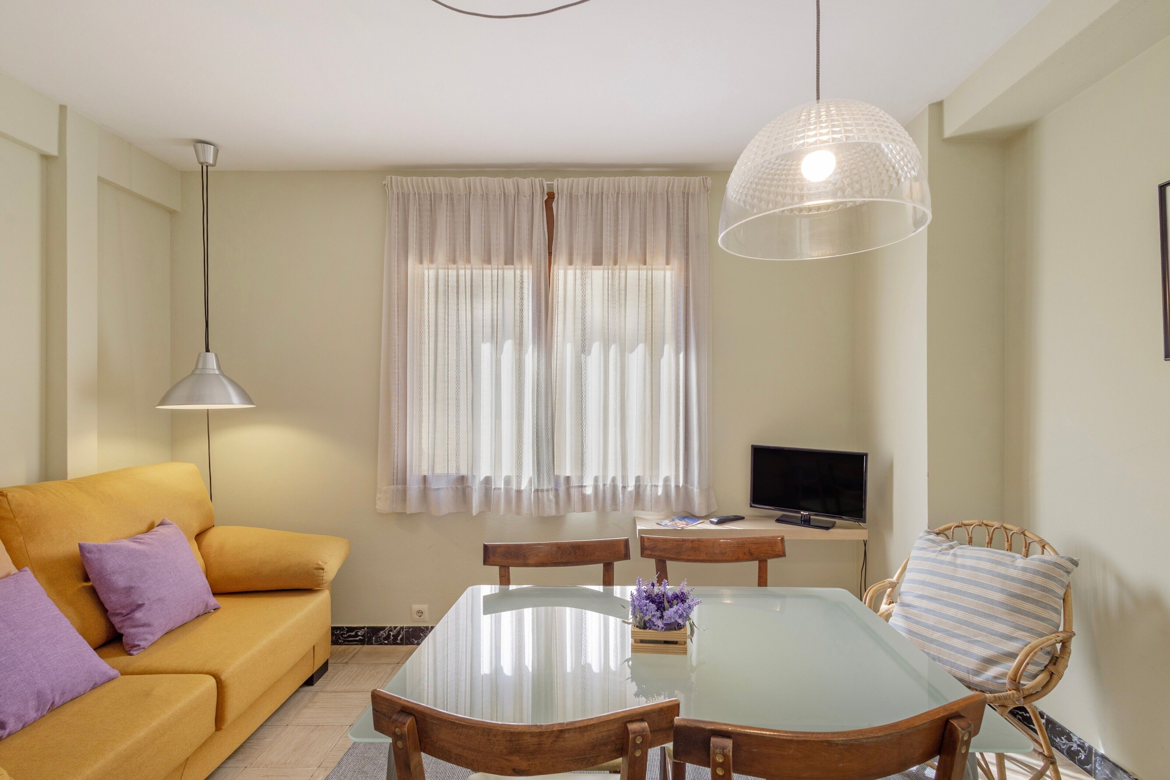 Una sala de estar con sofá, una mesa de comedor con superficie de vidrio, sillas, televisor y ventana con cortinas.