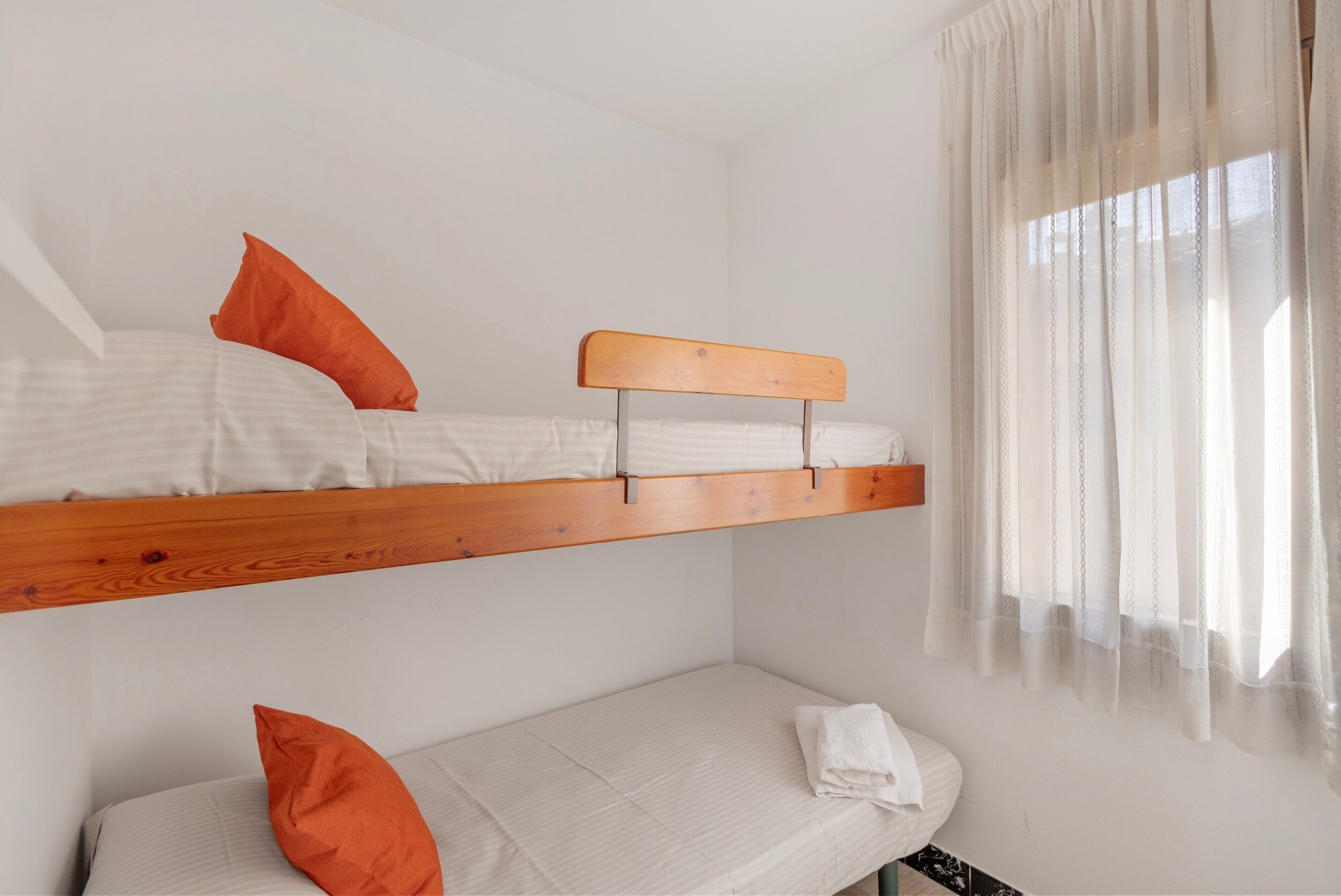 A bunk bed with a wooden headboard, a single bed below, and a window with sheer curtains.