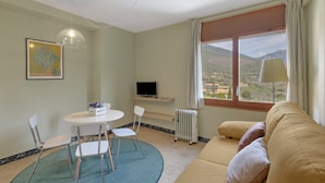 Een kamer met een bank, een eettafel met stoelen, een televisie, een radiator en een raam met uitzicht.