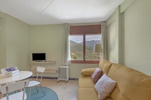 Una habitación con sofá, televisión, mesa y sillas, radiador y ventana con vistas a montañas.