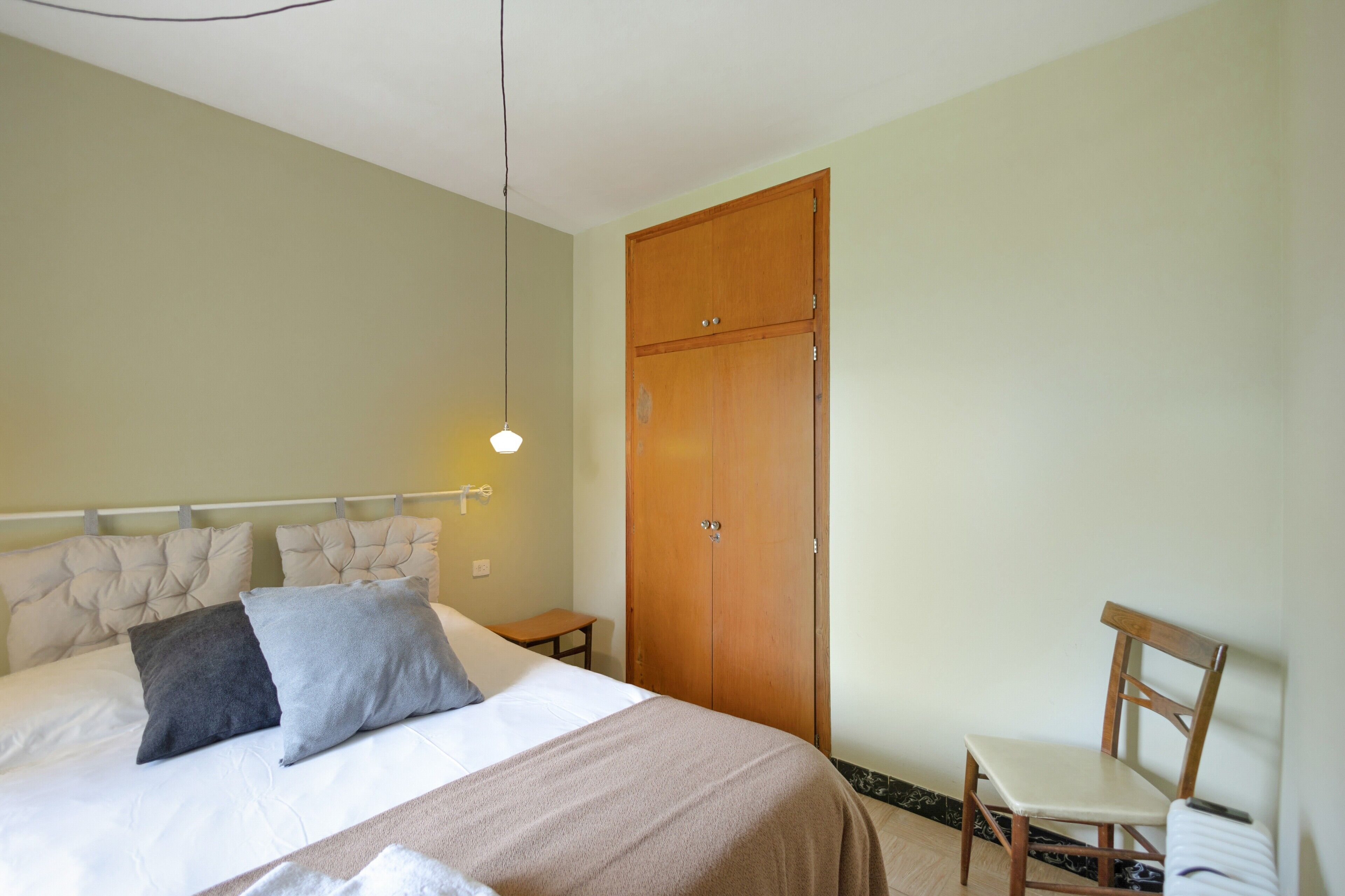 A bedroom with a bed, bedside table, chair, wardrobe, and a hanging light fixture.