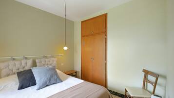 A bedroom with a bed, bedside table, chair, wardrobe, and a hanging light fixture.