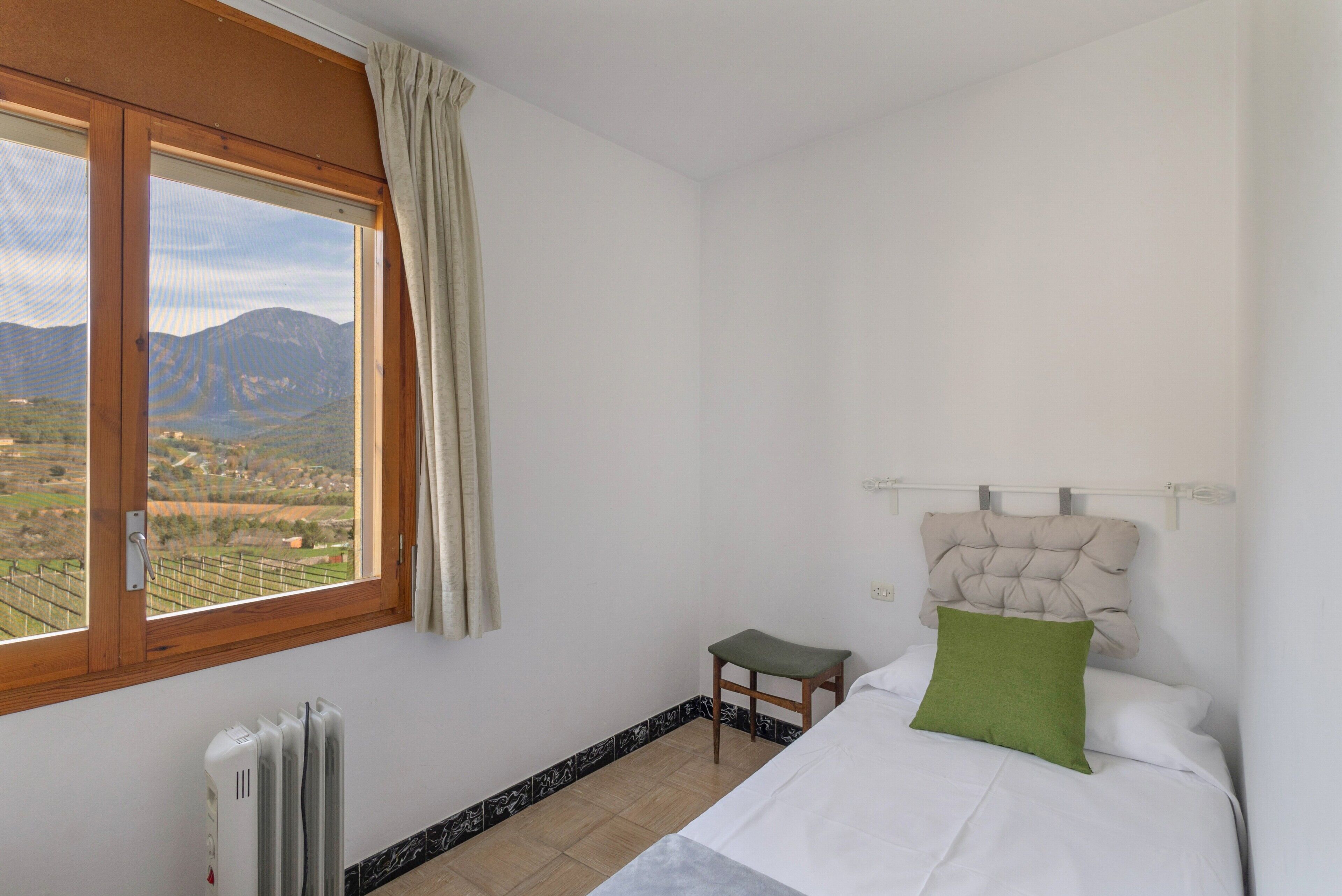 Un dormitorio con cama, un taburete pequeño, un radiador, una ventana con vistas a montañas y una cortina.