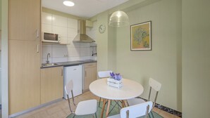 Een functionele keuken met een magnetron, spoelbak en kookplaat, een eettafel met stoelen en een wandklok.