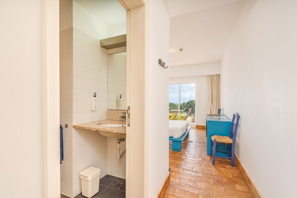 Un couloir mène à une salle de bain équipée d’un lavabo, de toilettes et d’une douche. On y trouve un lit avec une tête de lit bleue et une fenêtre avec des rideaux.