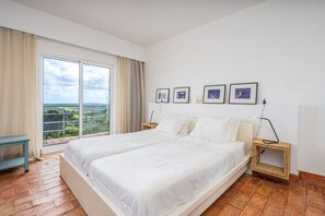 A bedroom with a large bed, a view of greenery through the window, and a small bedside table with a lamp.