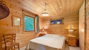 A wooden cabin room with a bed, a chair, a window, a lamp, and a painting on the wall.