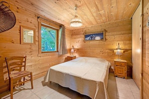 A wooden cabin room with a bed, a chair, a window, a lamp, and a painting on the wall.